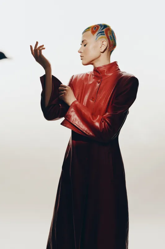 Creative woman with colorful shaved hairstyle wearing a shiny red leather coat, posing thoughtfully against a plain white background in a minimalist studio setting.
