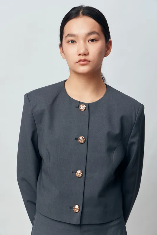 Portrait of a young woman in a gray formal jacket with decorative buttons standing against a plain white background. She has dark straight hair pulled back and a neutral expression.