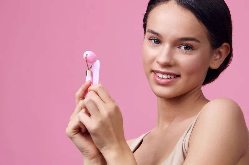 Young woman with natural makeup holding facial roller against a pink background, showcasing skincare routine and healthy lifestyle