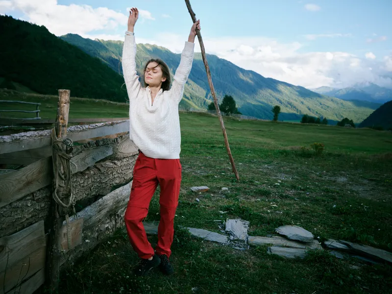 Mountain outdoor scene with a figure in a white sweater and red pants standing by a wooden fence, arms raised gripping a rope, expansive valley and clear sky