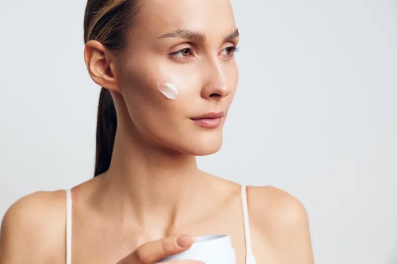 A young woman with clear skin applies cream to her cheek while holding a white jar. She looks thoughtfully to the side against a plain light background, emphasizing skincare and beauty routine.