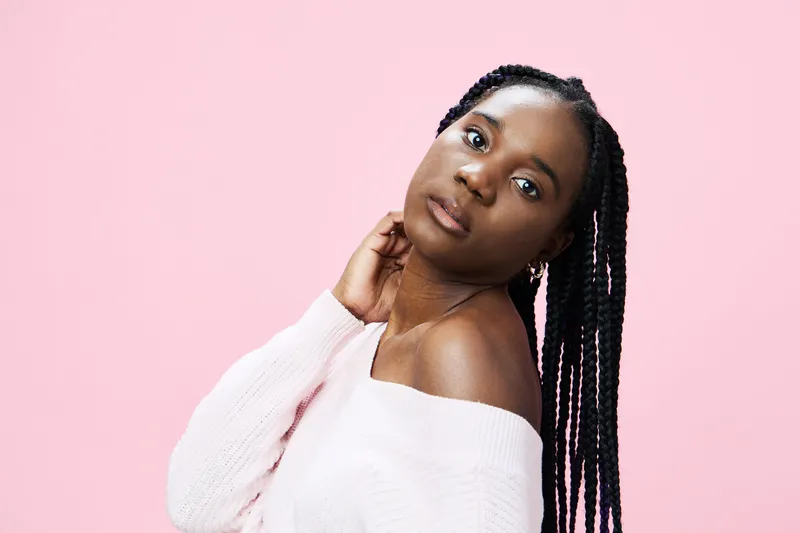 Portrait of a young Black woman with braided hair, wearing a soft pink off shoulder sweater, against a pastel pink background, conveying confidence and elegance