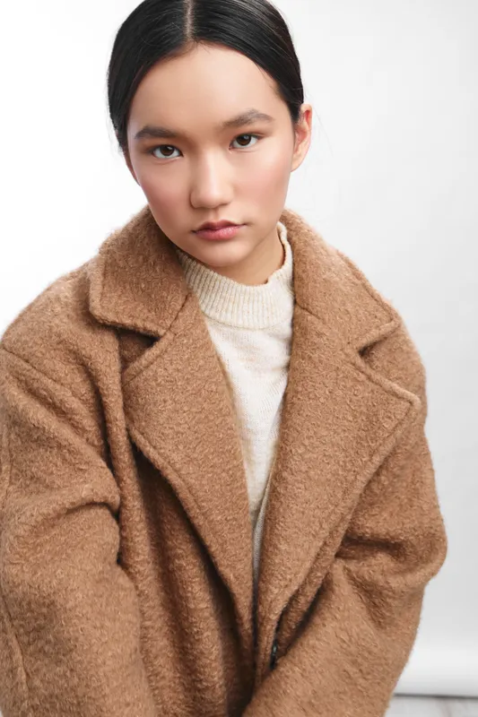 Asian woman with sleek dark hair posing in a soft studio setting. She wears a warm camel coat and light sweater, presenting a calm, confident, contemporary portrait.