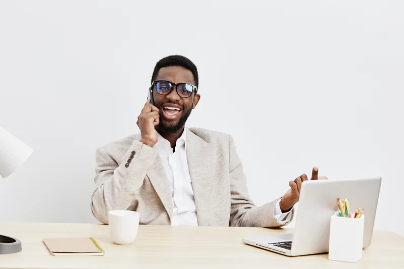 Happy smiling young man of African descent talking on mobile phone in modern office setting, wearing beige blazer and white shirt, working at desk with laptop and white background, lifestyle concept