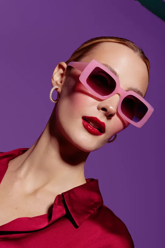 Stylish woman with pink sunglasses and red lipstick poses against purple background. Fashion portrait with bold colors and modern accessories. Concept of glamour and confidence.