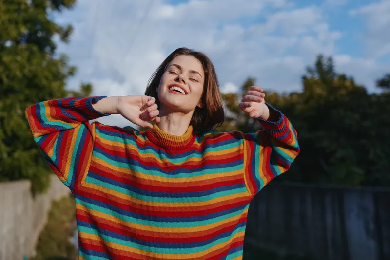 Young adult woman smiling in a colorful striped sweater outdoors, stretching arms with eyes closed, casual outfit and short brown hair in sunlit park for lifestyle, travel and wellness.