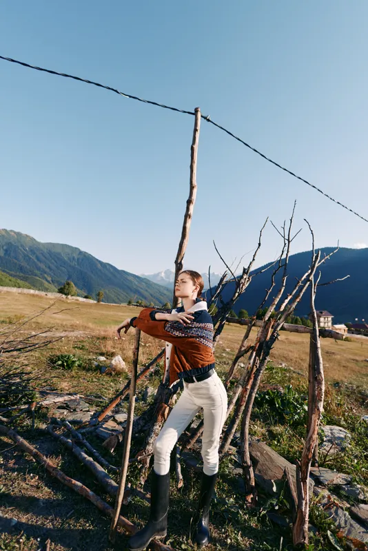 Woman in countryside by rustic fence and mountains, wearing patterned sweater and boots, leaning on wooden posts in rural landscape meadow under clear blue sky