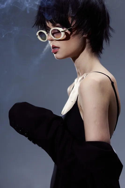 Stylish woman in modern sunglasses and black outfit exhaling smoke with moody lighting and dark background.