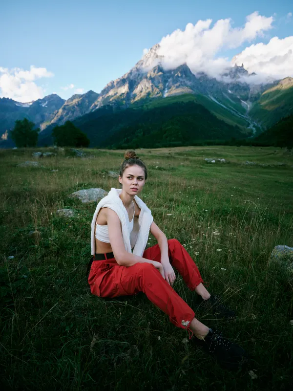 Woman sits in a meadow with red pants and white vest, set against dramatic alpine mountains and blue sky, conveying outdoor fashion, travel vibe, and peaceful nature