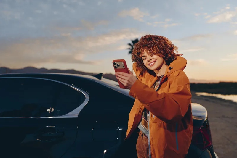 Woman lifestyle scene, smartphone selfie near car at sunset on open road, smiling person in orange jacket outdoors by parking lot, warm evening light, candid travel mood