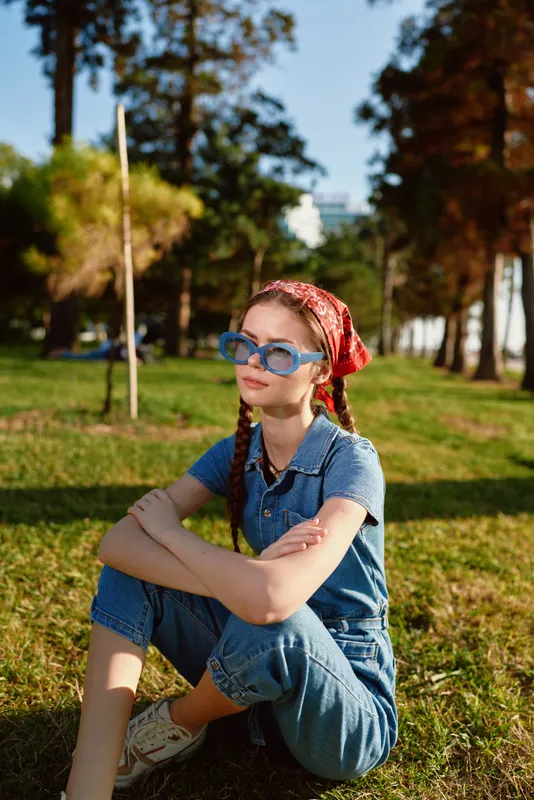 Lifestyle woman in modern boho-western denim street style sits outdoors wearing blue jean jumpsuit, red bandana, and blue sunglasses under warm sunlight.