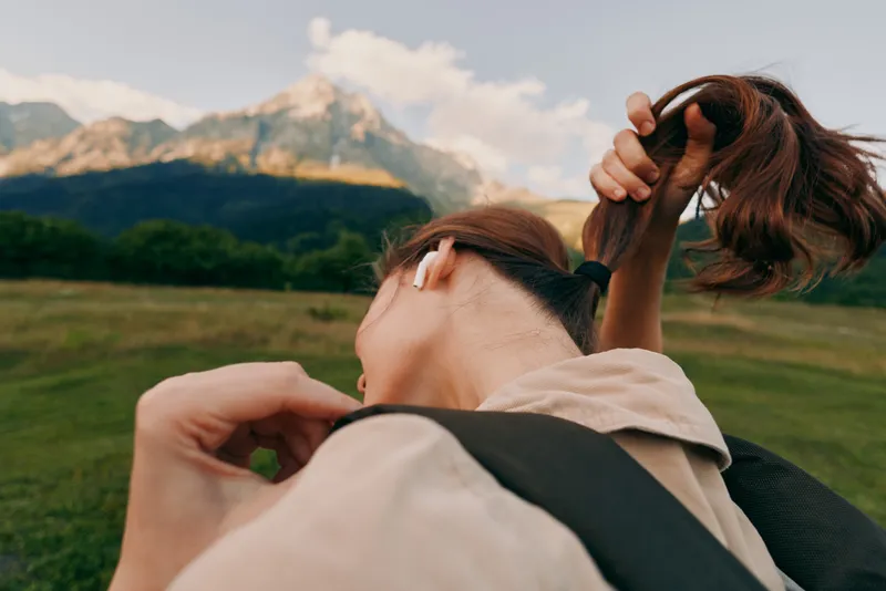 Young woman with a ponytail ties her hair back outdoors against a mountain landscape, a casual, carefree moment captured in daylight.