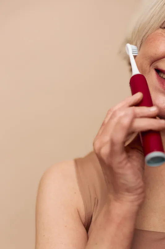 Close-up of a smiling mature woman using a red electric toothbrush on beige background. Concept for dental care, oral hygiene, and healthy lifestyle in senior adults.