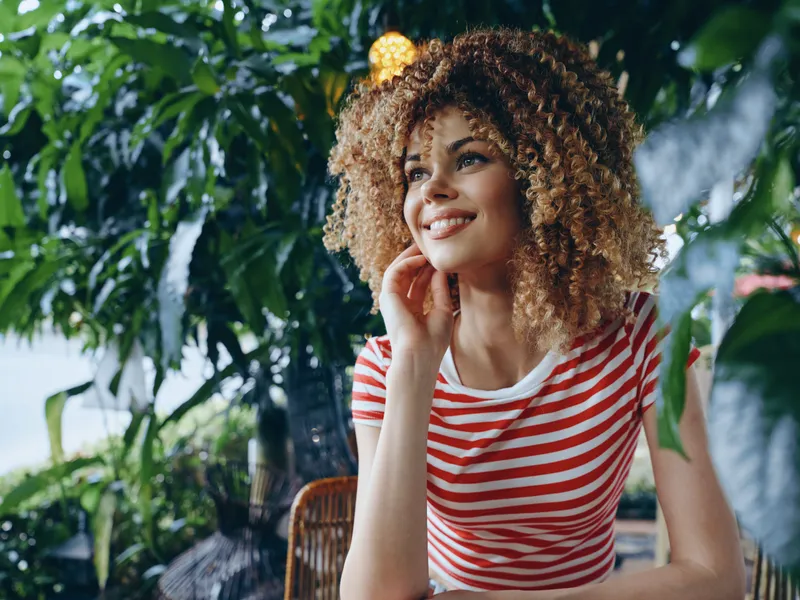 Portrait of a joyful woman with curly hair wearing a red and white striped shirt, posed outdoors among lush plants, warm light highlighting her smile.