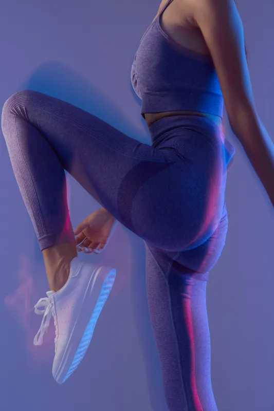 woman sport fitness wearing purple athletic leggings and top, stretching her leg in modern studio with blue and pink lighting, active lifestyle concept