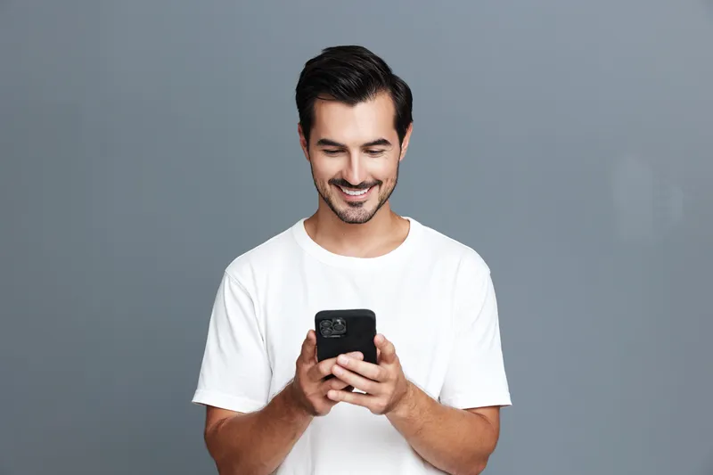 Smiling man using mobile phone, dressed in casual white t-shirt, standing against a solid grey background, showcasing modern lifestyle and technology connection.