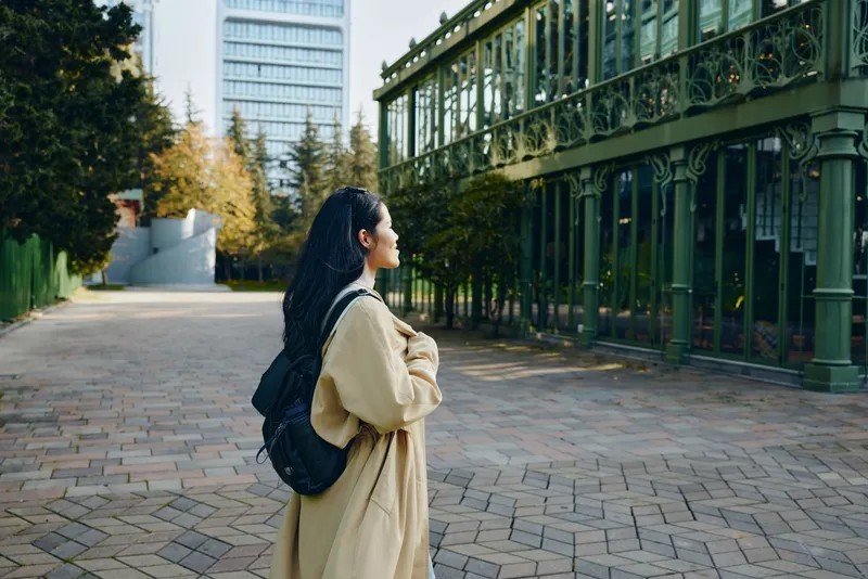 woman with backpack wearing a beige coat stands on a tree lined street admiring ornate green architecture in a calm urban setting during autumn, conveying exploration, style, and quiet elegance