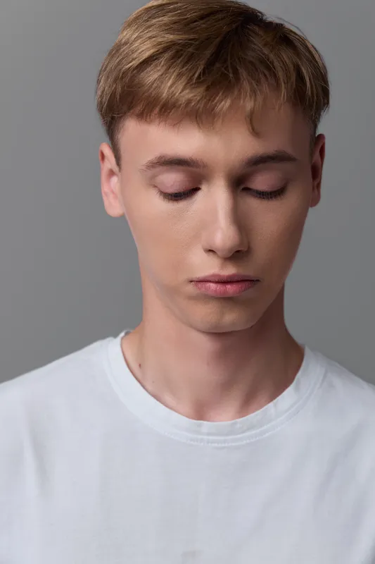 Young man with white tshirt and calm expression, head bowed in a minimalist studio with gray backdrop, creating a quiet mood suitable for fashion, lifestyle and indoor photography
