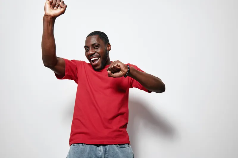 African American man with dark skin wearing red t shirt and jeans dancing happily with raised arm and smiling on white background. Joyful, casual and energetic expression.