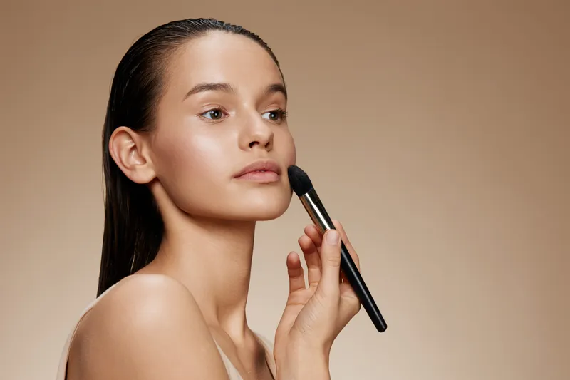 Beautiful young woman applying makeup with a brush, showcasing natural beauty and flawless skin tones on a soft beige background, emphasizing skincare and makeup artistry