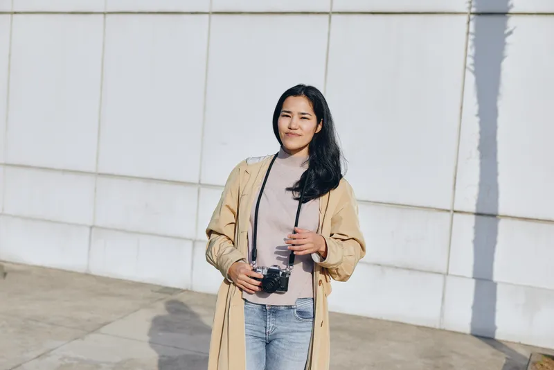 Woman with a camera stands outdoors in a casual beige jacket and denim jeans, smiling softly under bright daylight, ready for street photography and everyday storytelling