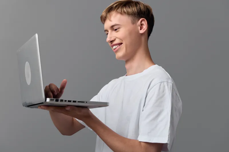 Casual young man with laptop, smiling while using a slim notebook computer in a simple studio setting, conveying remote work, tech hobby and everyday digital collaboration.