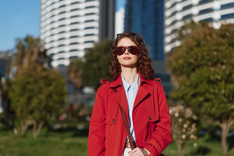 Woman in a red coat stands in a sunny urban park with modern buildings behind, wearing sunglasses and a light blue shirt, posing confidently.