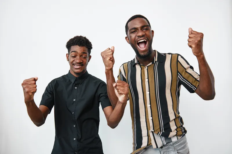 Two young black men with dark skin celebrating success with raised fists and happy expressions in casual clothing on white background. Friendship and joy concept.