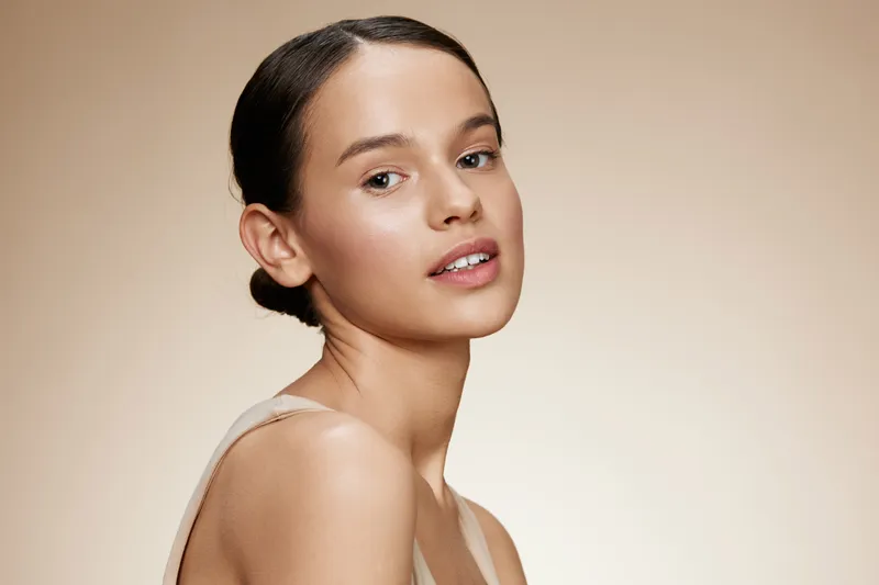 A young female model with a natural makeup look and a neutral background, showcasing effortless beauty and confidence with soft lighting and warm tones
