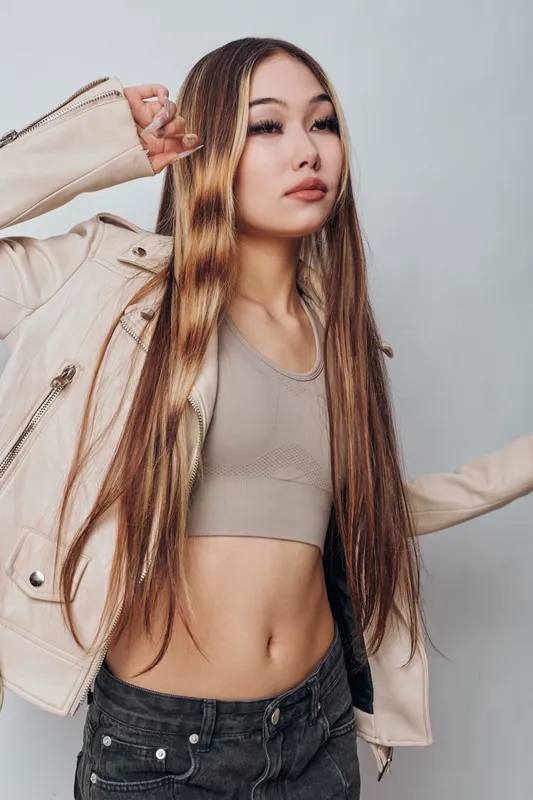 Young woman with long braided hair wearing beige crop top and light jacket posing confidently against plain background, fashion and style concept.