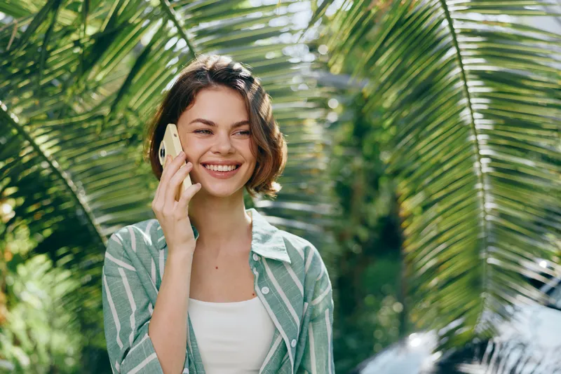 Young woman smiling while talking on a smartphone outdoors among lush palm leaves, sunny day, casual outfit.