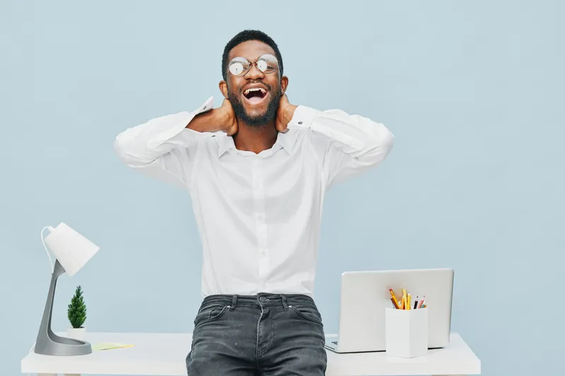 Vibrant, joyful man expressing excitement in bright office environment, wearing white shirt with glasses, sitting at desk with laptop, plant, and stationery, capturing a cheerful and energetic mood. P