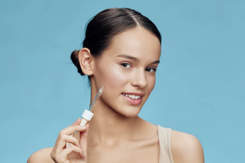 Young woman with brown hair holding a dropper with serum against a light blue background, showcasing healthy skin and beauty routine