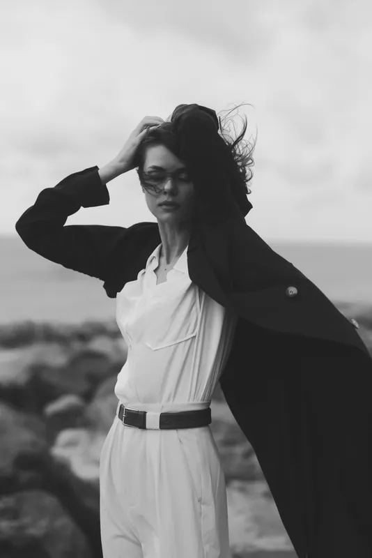 Fashionable woman on a windy coast wearing a white jumpsuit and dark coat, monochrome mood, dynamic movement, bold silhouette, cinematic style, confident and elegant