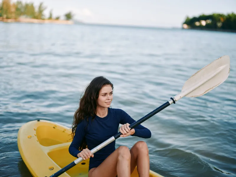 A woman paddling a bright yellow kayak glides across calm water, wearing a dark blue top and a focused expression as she grips a long paddle and concentrates on her stroke.