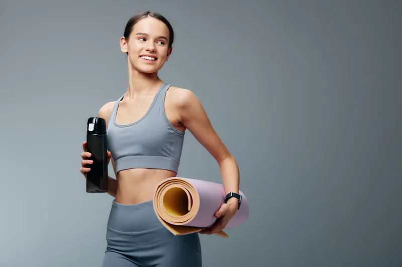 Fitness woman, athletic and confident, holds a water bottle and rolled yoga mat before a workout. She wears sportswear and stands ready for training in a clean studio.