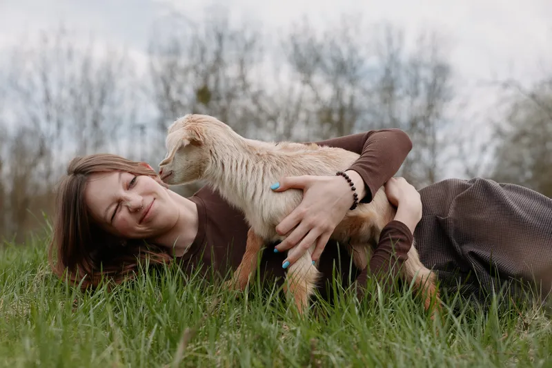 Young woman lies on green grass gently holding a baby goat while smiling with closed eyes in a natural outdoor setting during daytime spring season