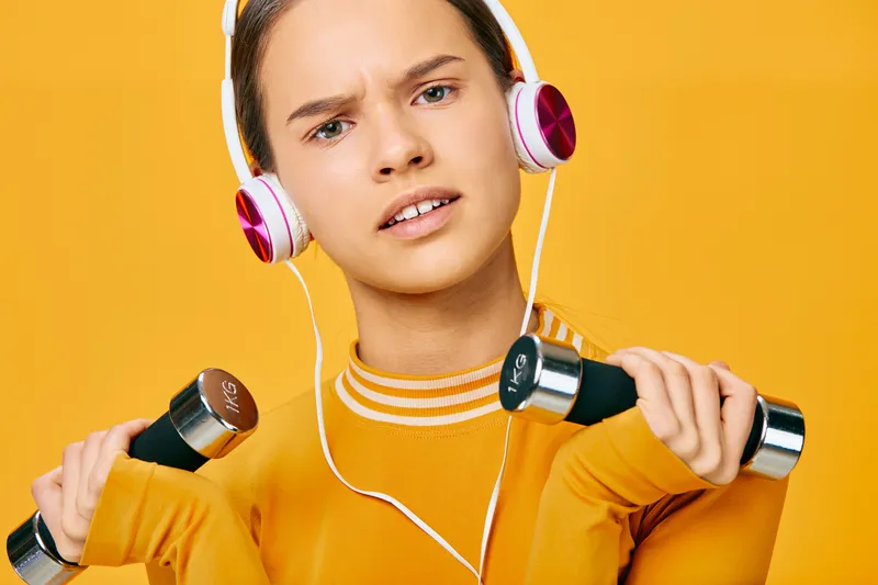Energetic woman in an orange sweater lifts dumbbells with headphones, showcasing fitness, strength training, and concentration against a vibrant yellow background.