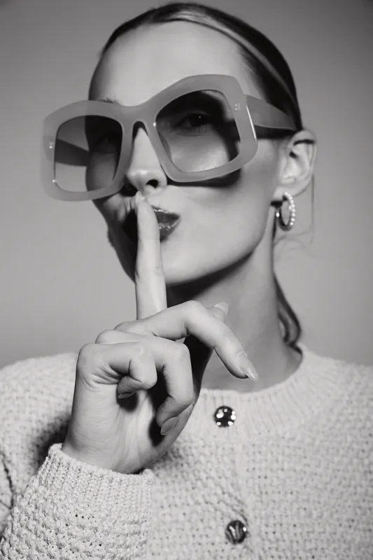 Stylish caucasian woman with pale skin wears large sunglasses and holds finger to lips in shushing gesture. Black and white portrait with fashionable mood and concept of secrecy.