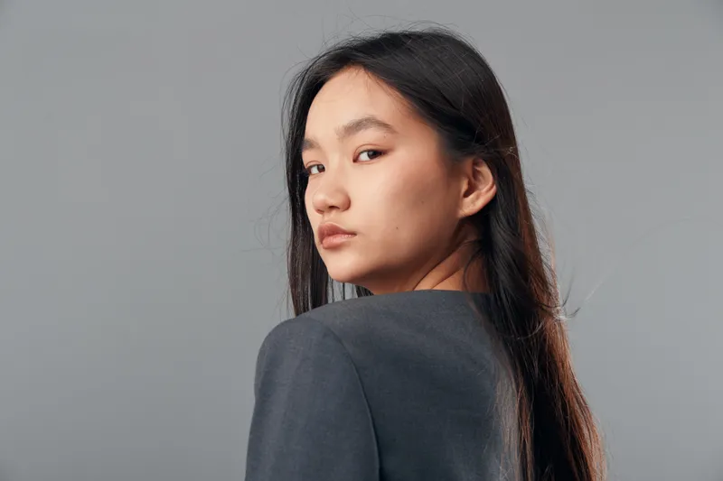 Portrait of young woman with long dark hair looking over shoulder with serious expression against plain gray background. Asian female model with natural makeup wearing gray top in studio.