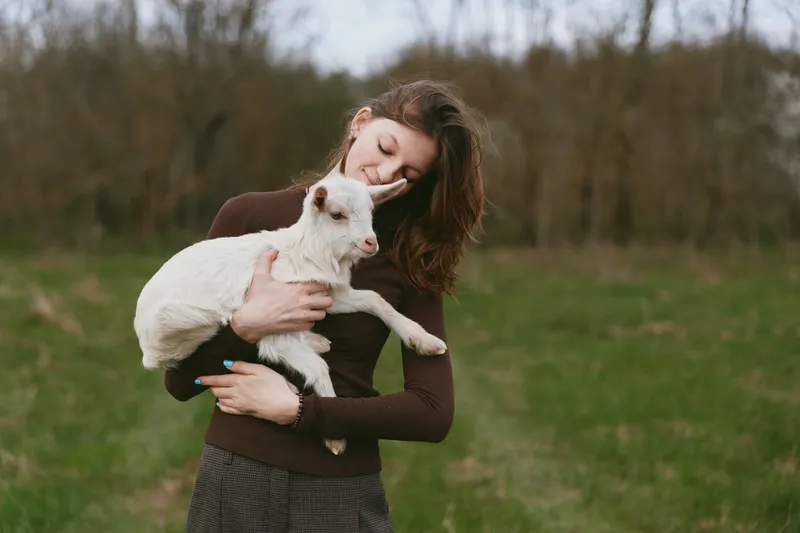 A woman in a brown sweater cradles a white goat kid in a sunlit meadow, smiling softly as they share a quiet moment. A lifestyle scene of animal bonding and care.