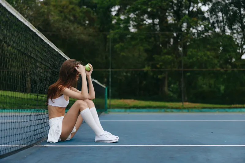 Young female tennis player in white sportswear sitting on a court, feeling frustrated with a tennis ball, surrounded by greenery, capturing the essence of sports emotion