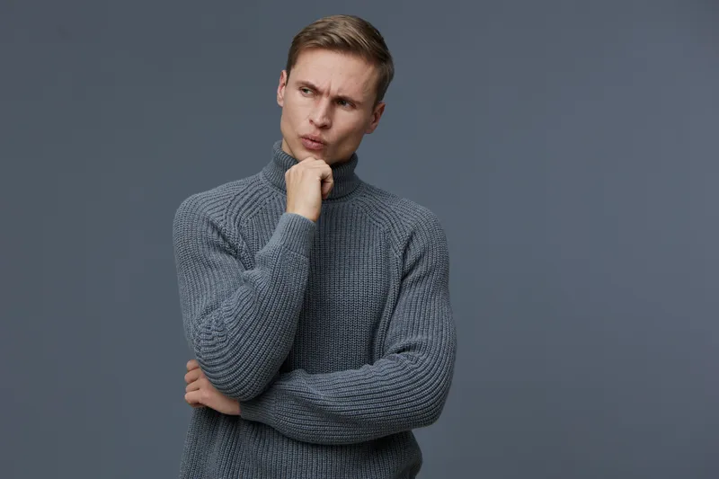 Thoughtful young man with light skin and short hair, wearing a cozy gray turtleneck sweater, looks contemplative with hand on chin, isolated on a plain dark gray background. Concept: pondering and cur