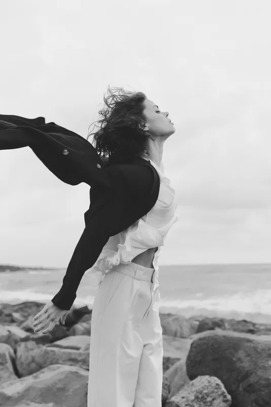 Woman stands on a rocky shoreline with wind sweeping her jacket, a fashion forward pose by the sea that captures freedom, movement and a moody monochrome mood