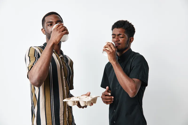 Two young black men with dark skin drinking coffee from white cups in casual clothing on white background. Friends enjoying hot beverage and social moment together indoors.