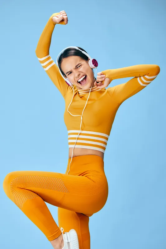 Fitness woman in a bright orange athletic outfit dances energetically with headphones on, against a blue background, capturing dynamic movement, joy, and fitness in a vibrant workout moment.
