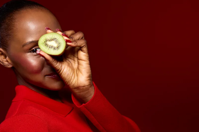 Smiling African woman with dark skin holds half of kiwi fruit near eye. She wears red sweater and poses on red background. Concept of healthy lifestyle, fresh food, and natural beauty.