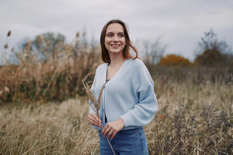 Young woman stands in a windswept meadow smiling naturally while holding dried grass, conveying authenticity and a genuine outdoor lifestyle in soft overcast autumn light.