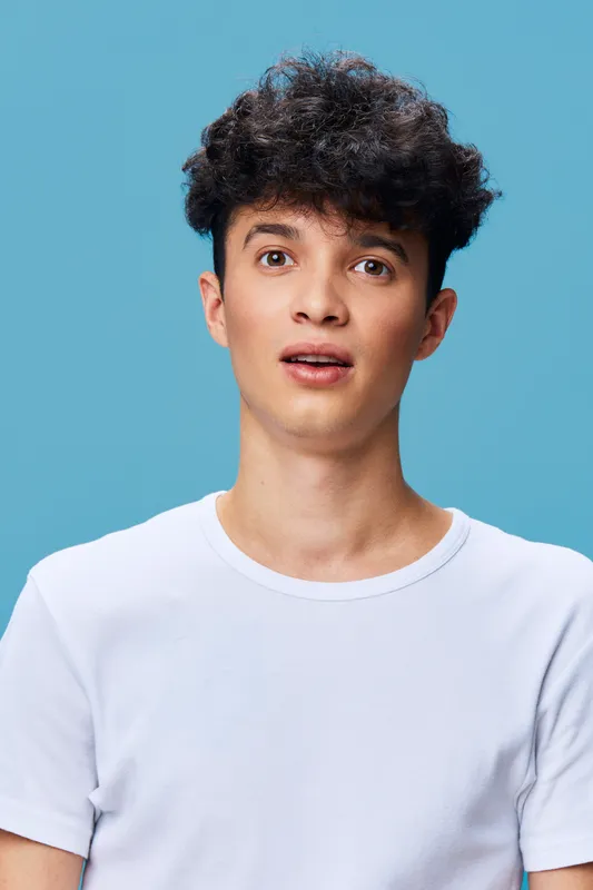 surprised young man with curly hair wearing white t-shirt looks amazed against blue background, expressing shock and curiosity, casual style portrait in bright light.