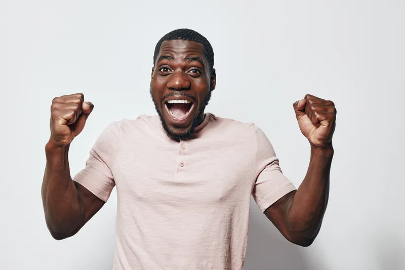 Excited African American man with dark skin wearing light pink shirt celebrating success with raised fists and wide open mouth on white background. Joyful expression and casual style.